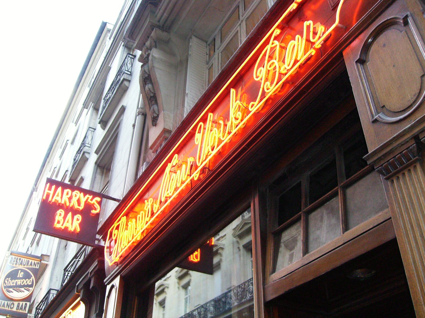 Неоновая вывеска Harry's New York Bar красным курсивом над деревянным фасадом парижского бара, рядом прямоугольная вывеска HARRY'S BAR