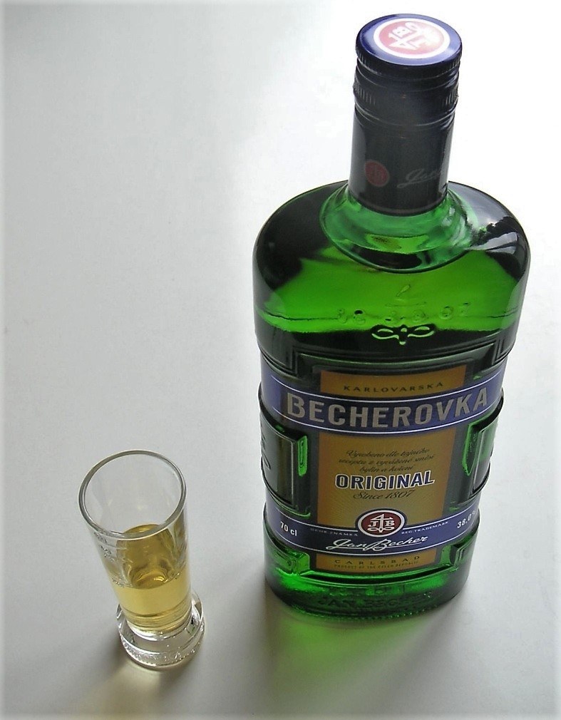 Зелёная плоская бутылка Бехеровки с золотисто-синей этикеткой и рюмка с янтарным ликёром рядом на светлом фоне