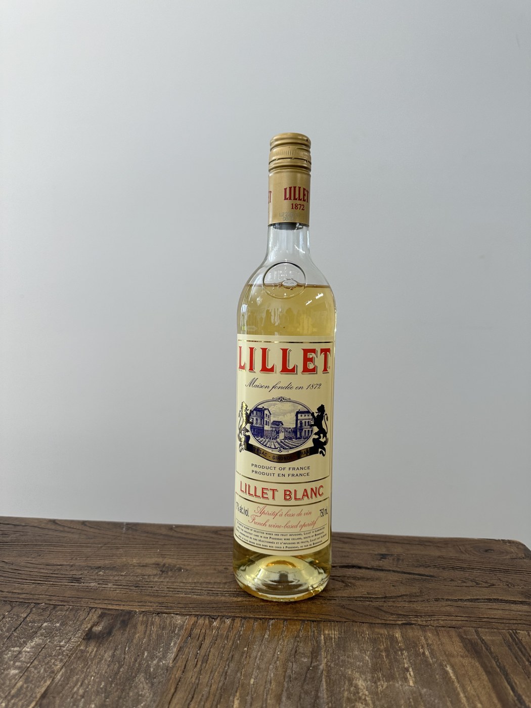 Бутылка светлого аперитива с кремовой этикеткой и красной надписью LILLET, с золотистым напитком внутри, стоит на деревянной столешнице на светлом фоне