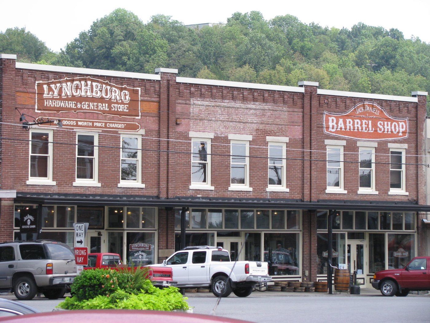 Кирпичный ряд домов на площади городка: вывески «LYNCHBURG HARDWARE & GENERAL STORE» и «BARREL SHOP», перед зданиями припаркованы пикапы, на заднем плане лесистый холм